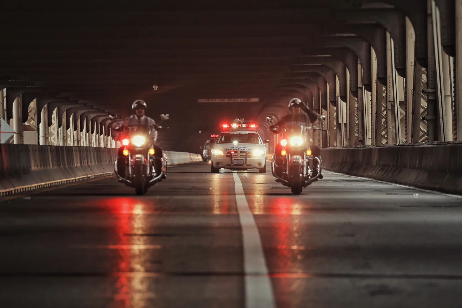 Eskorte bei Nacht — Foto: Norbert Banhalmi Polizeimotorräder mit roten Lichtern nähern sich nachts auf leerer Straße unter einer Kolonnade.