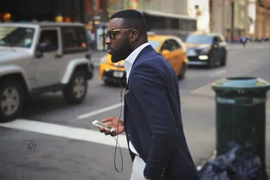 Man on a New York street — Photo: Norbert Banhalmi A man in a navy suit and white shirt walks a Manhattan street with headphones in, a yellow cab passing behind him.