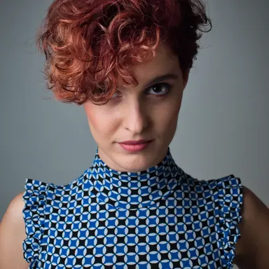 Portrait with red curls — Photo: Norbert Banhalmi Studio portrait of a woman with short red curls in a blue and white geometric top, calm gaze into the lens.