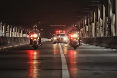 Escort at night — Photo: Norbert Banhalmi Police motorcycles with red lights approach along an empty road beneath a colonnade at night.