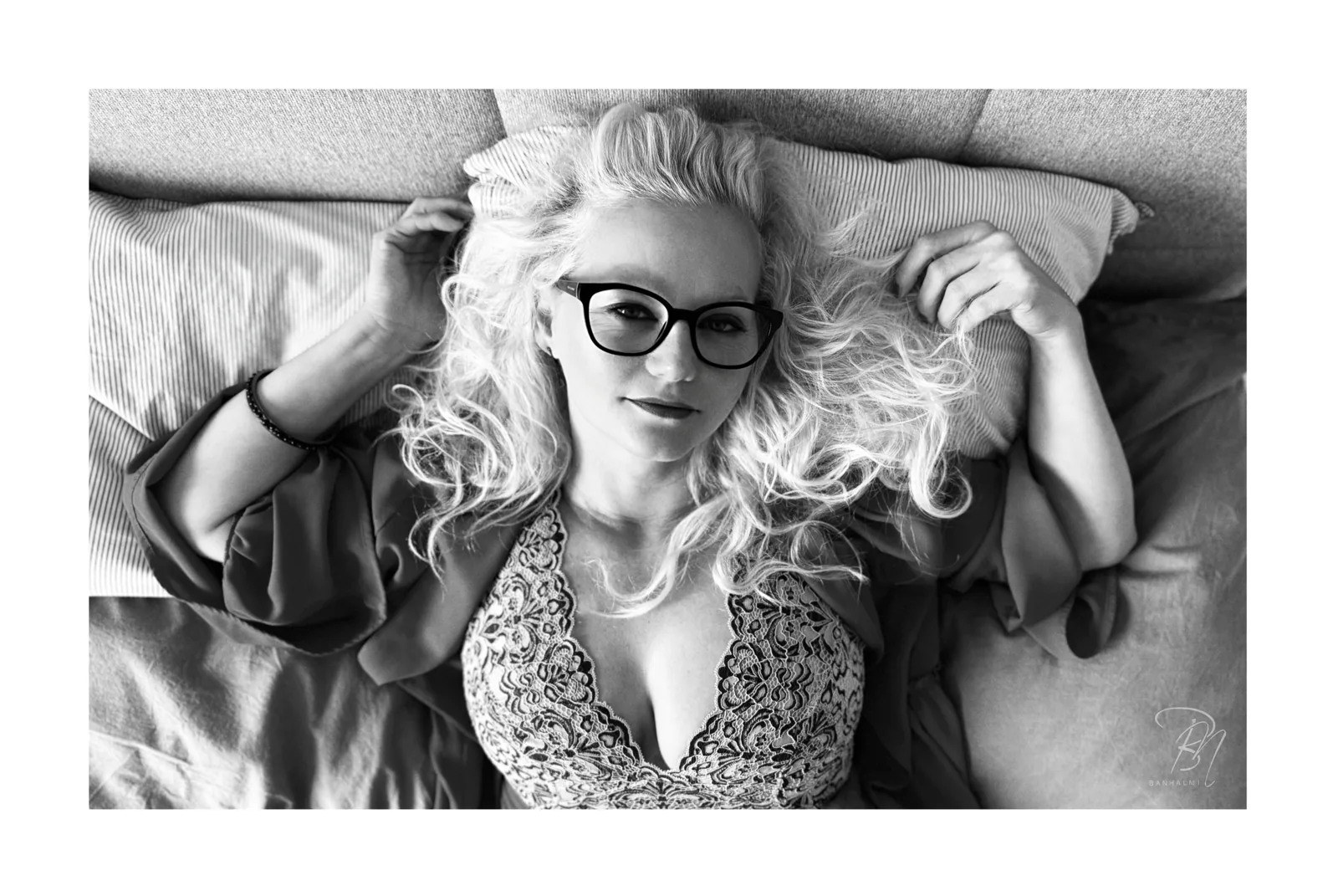 Im Licht liegend — Foto: Norbert Banhalmi Eine Frau mit hellen Locken und dunkler Brille liegt auf einem Bett, die Arme über dem Kopf.