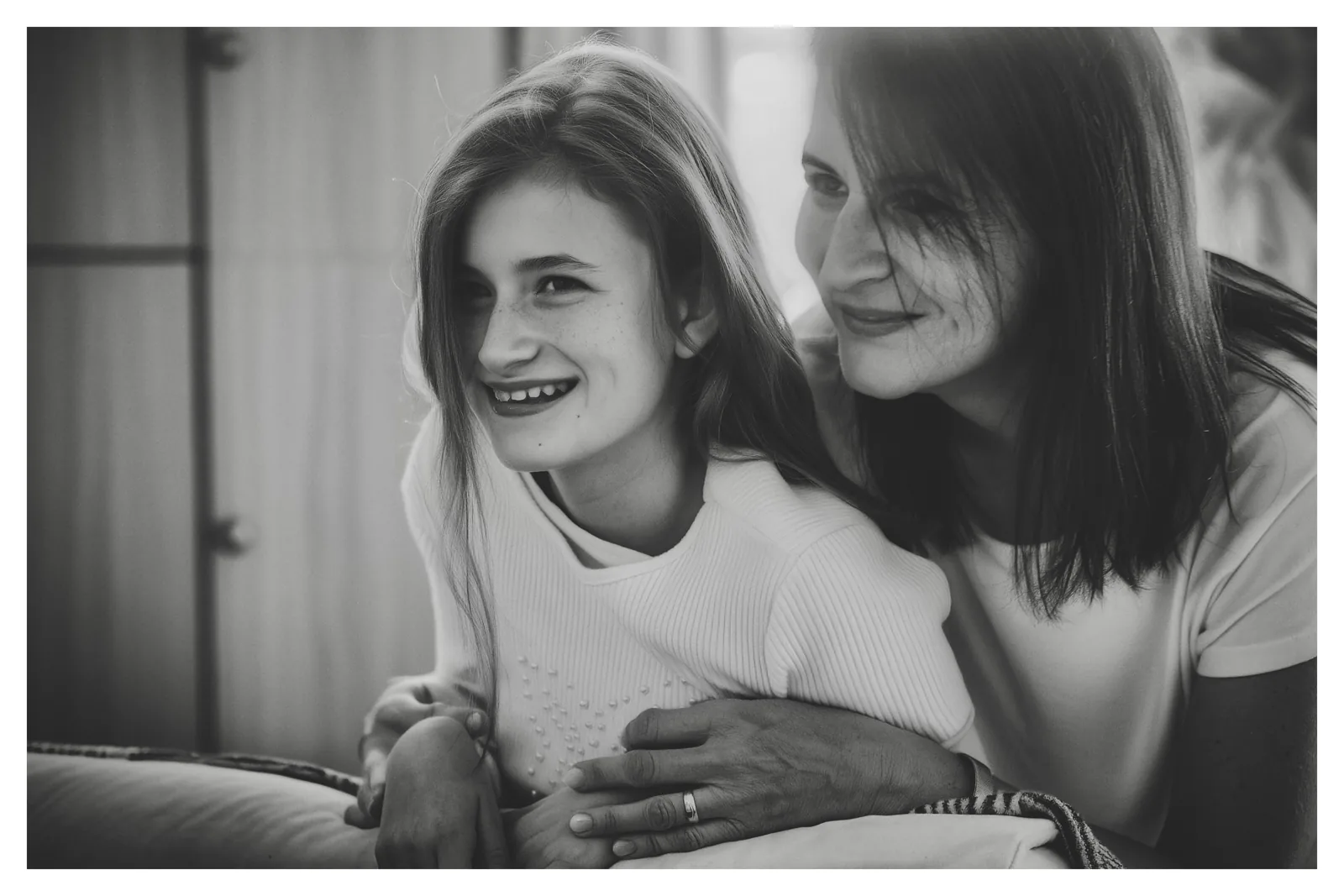 From behind the shoulder — Photo: Norbert Banhalmi A girl smiles into the room while a woman leans over her shoulder, hands resting in her lap.
