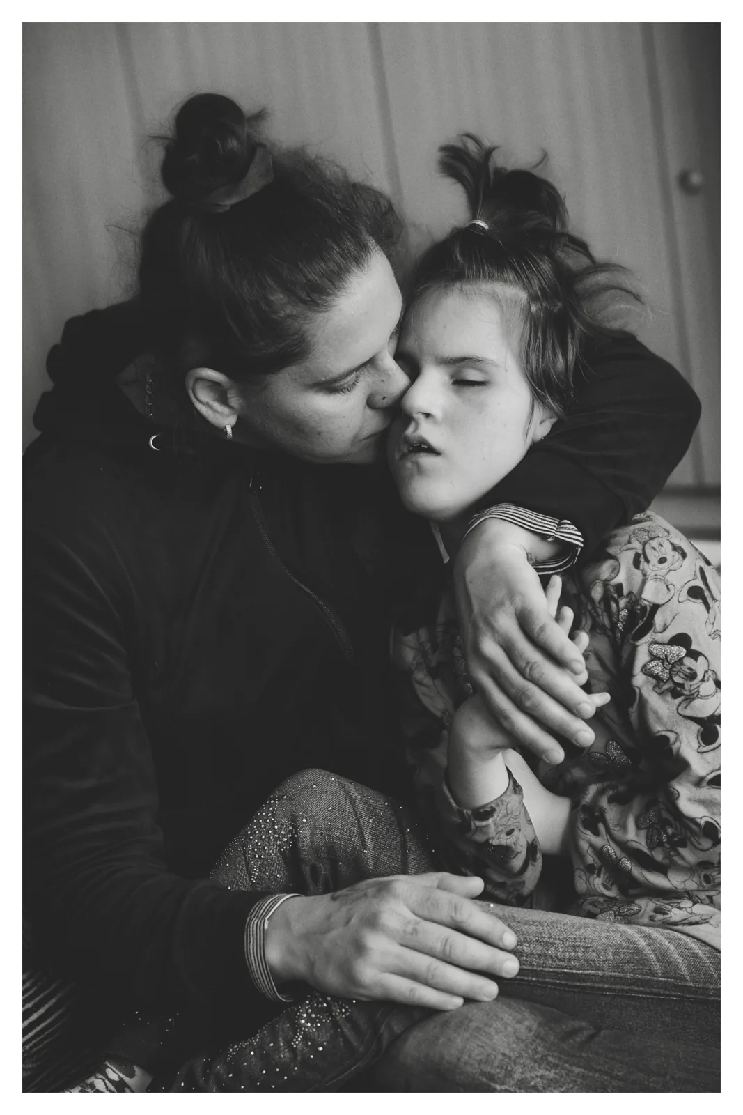 On her lap — Photo: Norbert Banhalmi A woman holds a girl on her lap and kisses her cheek.